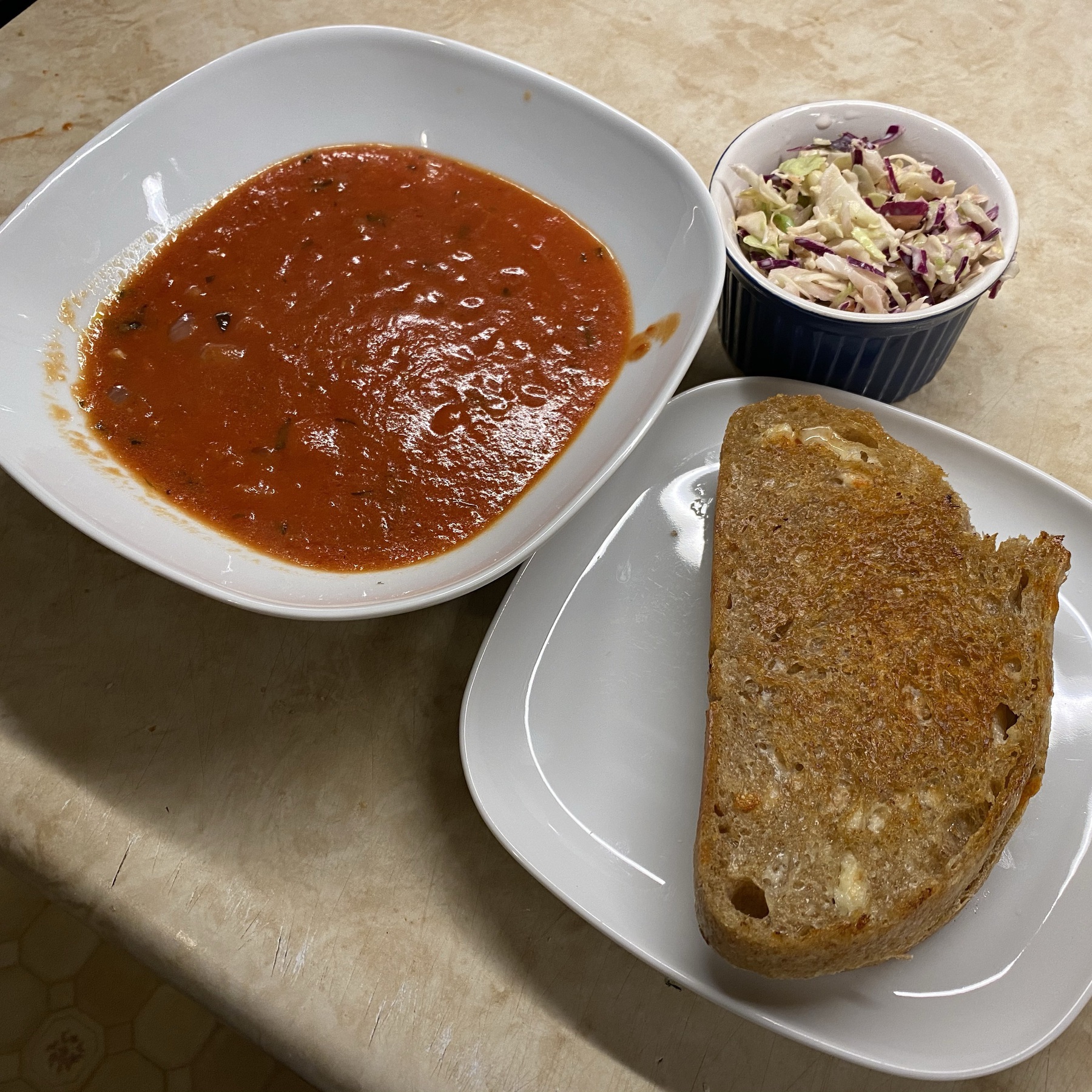 Bowl of tomato soup, grilled cheese sandwich on plate, and bowl of coleslaw.